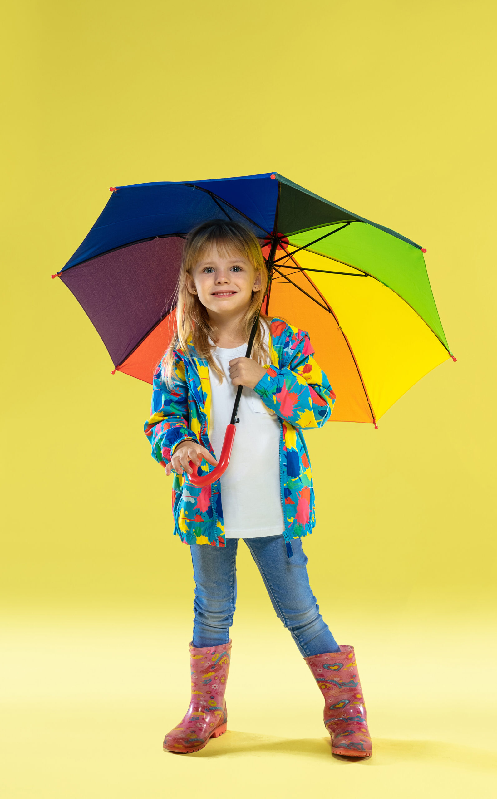 a full length portrait of a bright fashionable girl in a raincoat holding an umbrella of rainbow colors on yellow studio background scaled 1